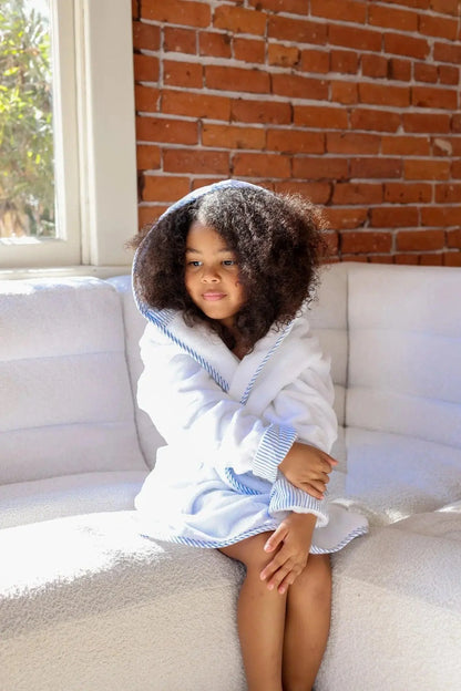 Child wearing a white hooded robe sitting on a white couch with a brick wall background