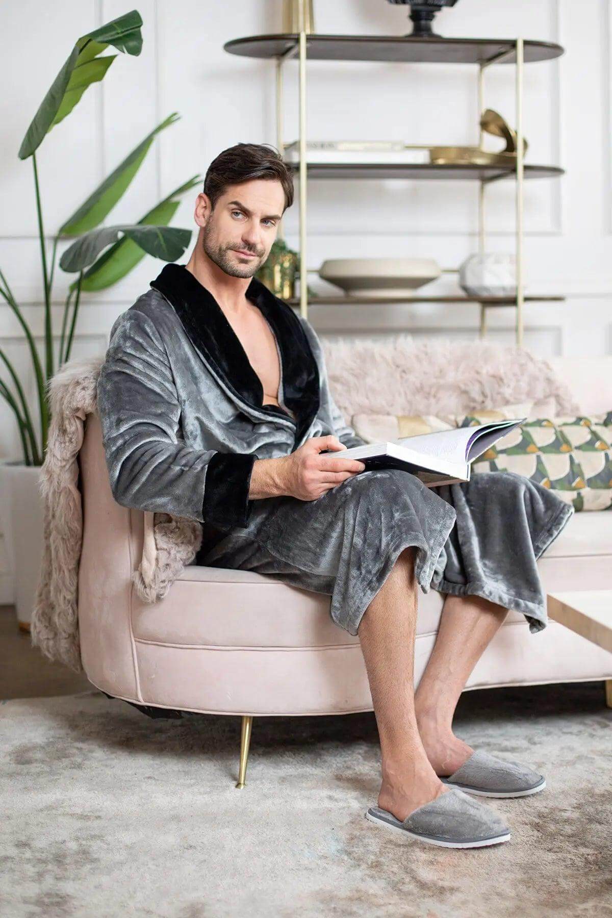 Man in a gray velvet robe sitting on a pink chair reading a book in a stylish room.