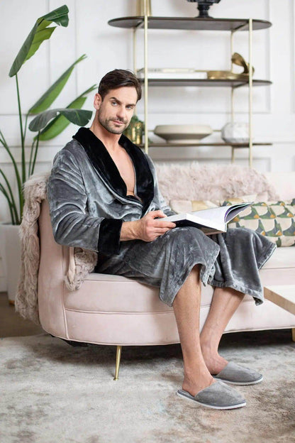 Man in a gray velvet robe sitting on a pink chair reading a book in a stylish room.