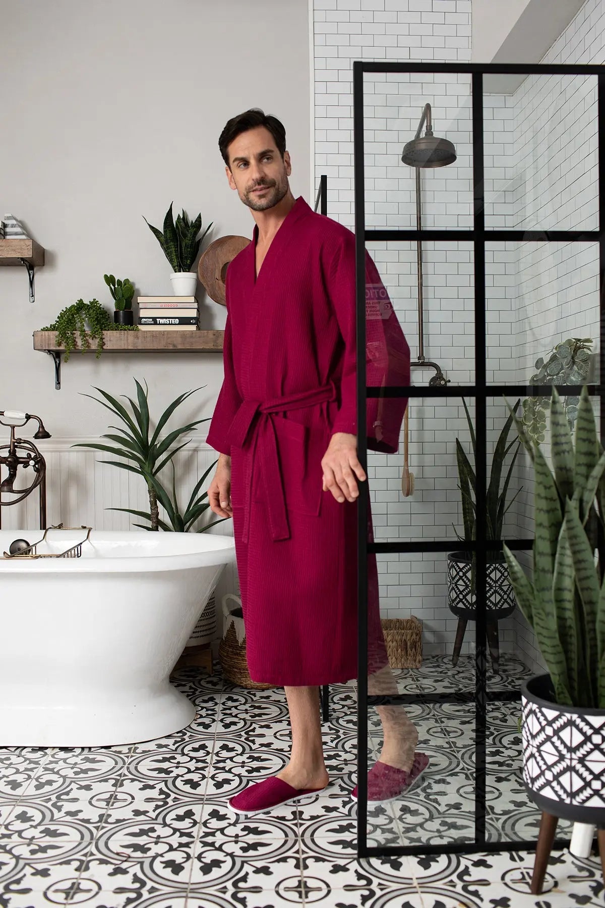 Man wearing a red robe standing in a bathroom with plants and a shower.